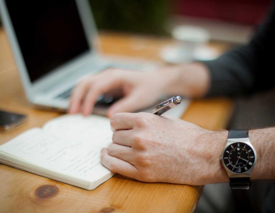 Hands writing decision notes in a notebook beside a laptop